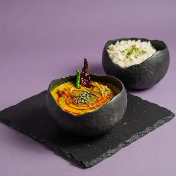 A black bowl of yellow dal with red and green chilies, beside a bowl of white rice.