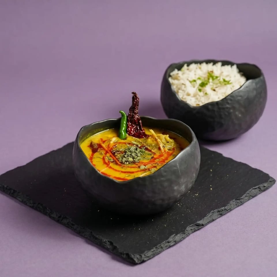 A black bowl of yellow dal with red and green chilies, beside a bowl of white rice.