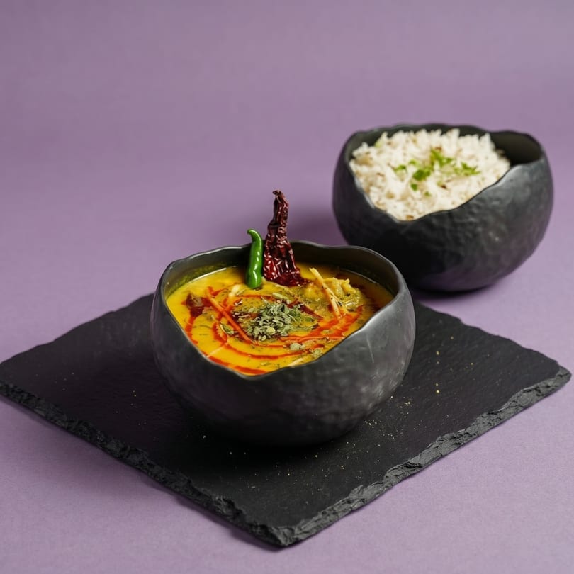 A black bowl of yellow curry with red and green chilies, next to a bowl of rice.