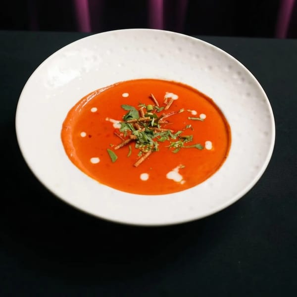 Vibrant red tomato soup with cream swirls, fresh herbs, and crispy croutons in a white bowl.