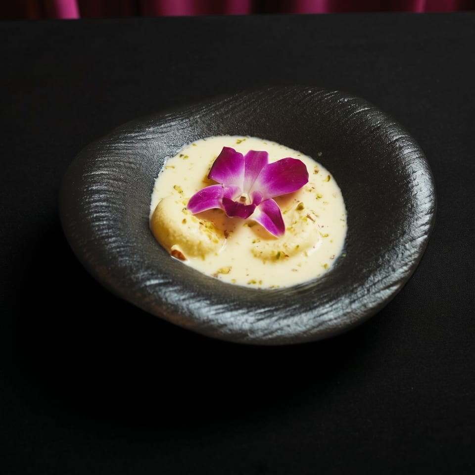 Rasmalai dessert in a black bowl, garnished with a purple orchid and pistachio crumbles.