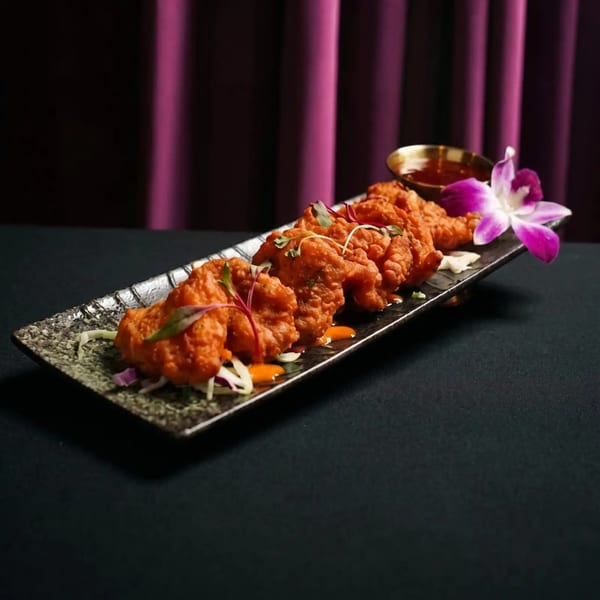 Crispy fried chicken pieces with sauce and an orchid on a long plate, against a purple curtain.