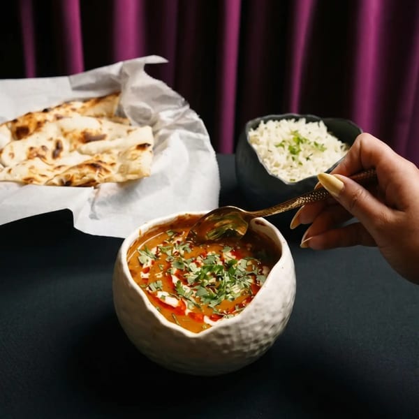 A hand spoons creamy Indian curry from a white bowl, with naan and rice in the background.