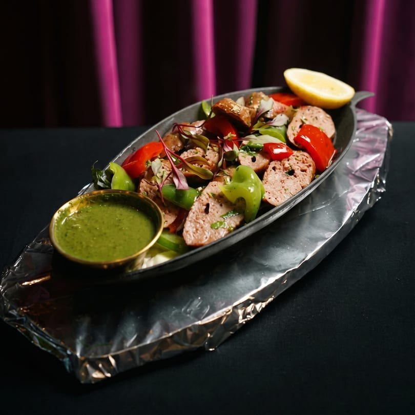 Sausage slices with red and green peppers, garnished with greens and a lemon wedge, served with green sauce.