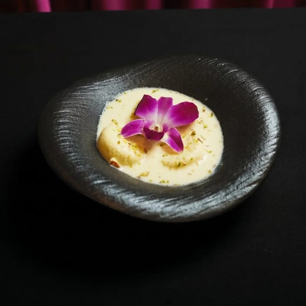 Creamy dessert with nuts and a purple orchid in a dark, textured bowl on a black surface.