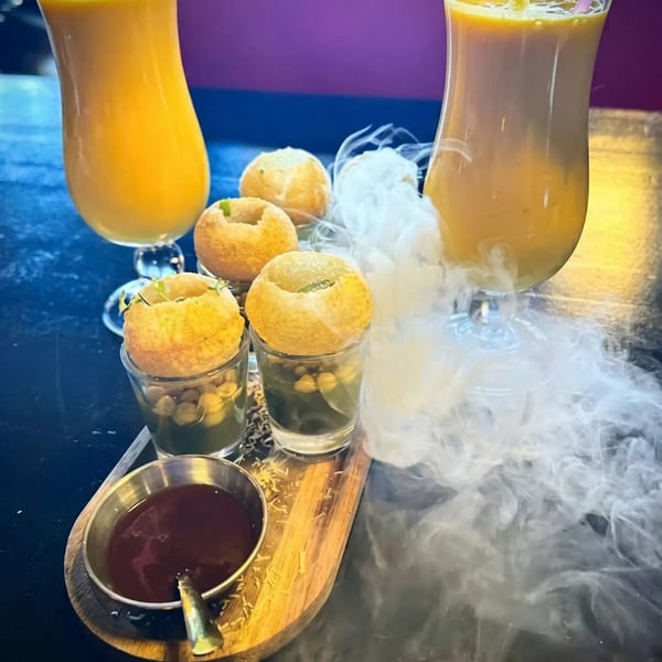 Pani Puri shots with two mango lassi drinks, served on a wooden tray with dramatic smoke.